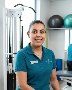 Smiling female physiotherapist in teal polo shirt standing in a modern gym with exercise equipment and fitness balls in background