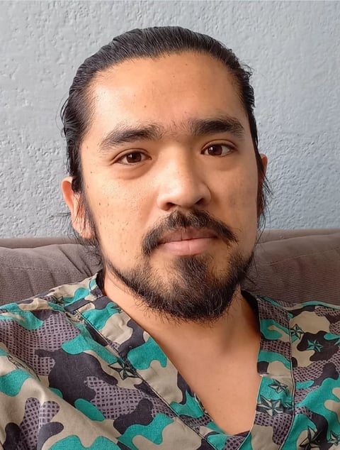 Portrait of a man with long dark hair and facial hair wearing a teal and purple camouflage patterned shirt against a white wall.