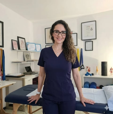Woman with glasses wearing dark scrubs in a clinical office with shelves of medical equipment and framed certificates on walls